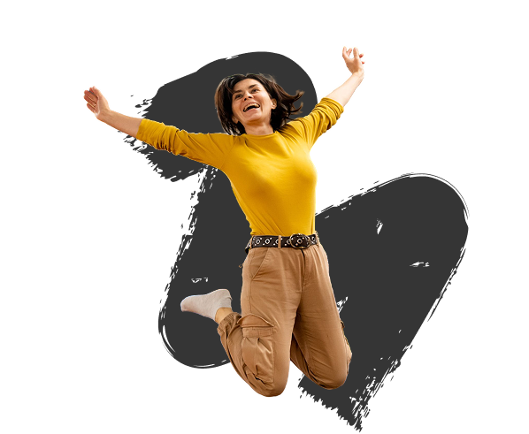 A woman in a yellow sweater and beige pants joyfully jumps in the air against a black background with a gray paint splash, conveying excitement.